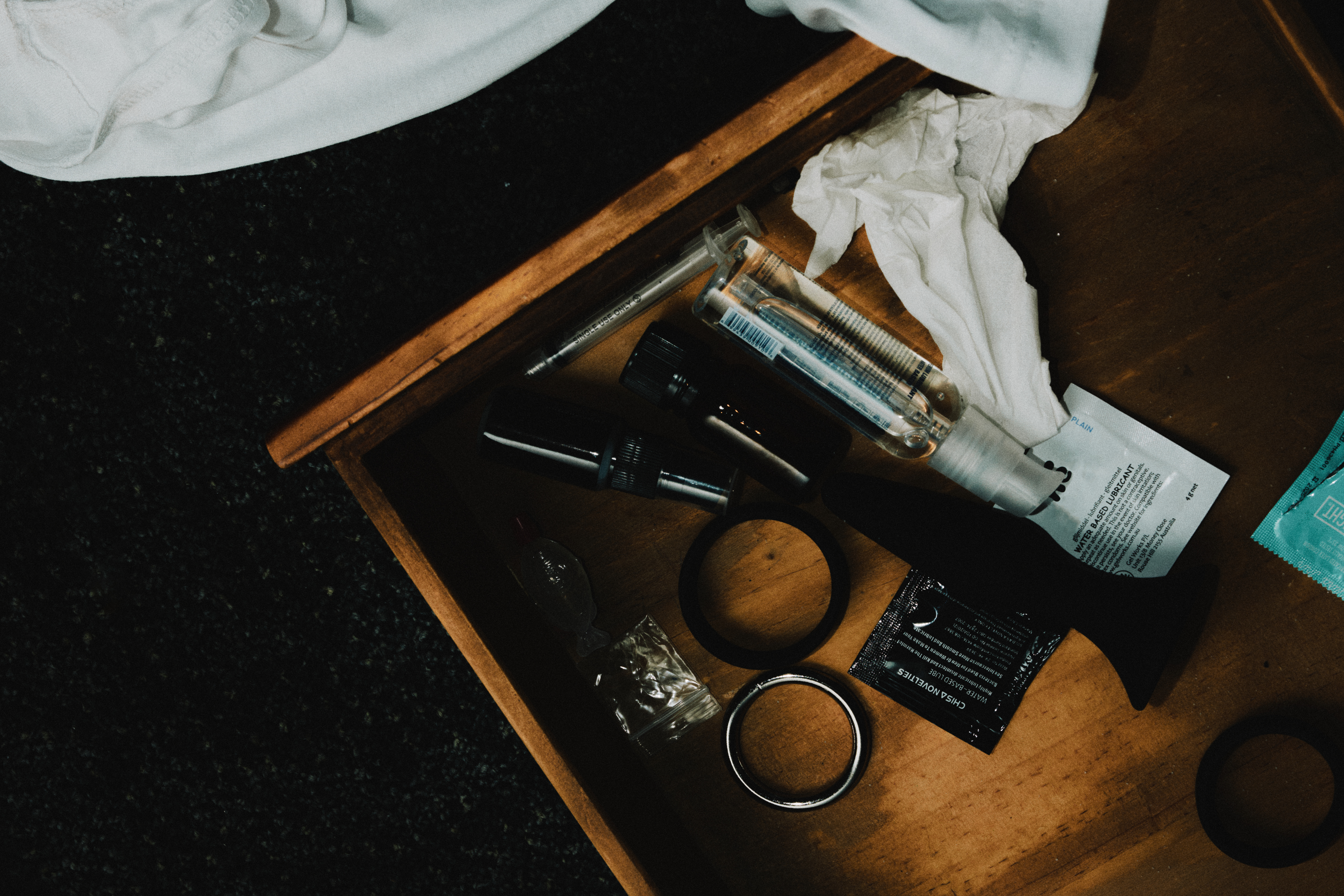 A small drawer beside a messy bed including a collection of sex and drug-related paraphernalia, including condoms, cock rings, lube, tissues, and a syringe.