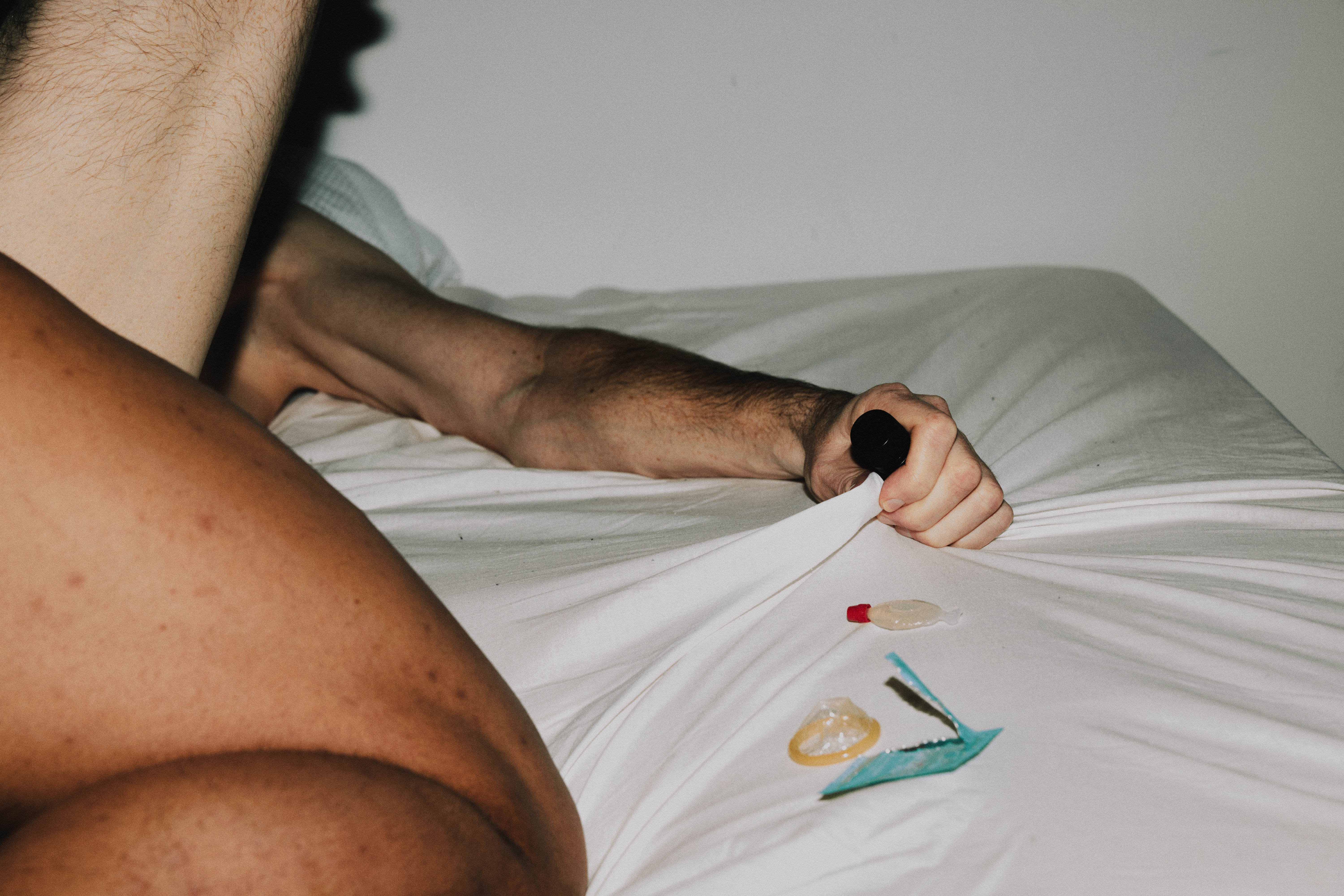 A tangle of male limbs on a white sheet in the throes of passion, with one clutching a poppers bottle beside an open lube packet and a container of G.